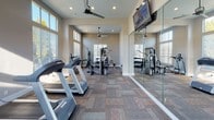 This single-floor fitness center is thoughtfully designed to support a comprehensive workout experience. The space is defined by soaring ceilings and an abundance of natural light streaming in through large, multi-pane windows positioned at both standard and clerestory heights, creating a bright and energizing atmosphere throughout the day. Warm terracotta-toned accent walls contrast with neutral gray tones to give the room a dynamic yet welcoming character.

The main workout floor is finished with durable, patterned carpet tiles in a multi-tone earthy palette, providing both comfort and practicality underfoot. Ceiling fans are strategically placed throughout to maintain airflow during intense workouts. Wall-mounted flat-screen televisions offer entertainment while exercising.

The cardio section is well-stocked with Precor treadmills and upright stationary bikes, all arranged to face the windows and mirrors for an open, spacious feel. A dedicated strength training area features a multi-station cable machine along with a free-weight rack and adjustable workout bench. Full-length mirrors line one wall, visually expanding the space and allowing users to monitor their form. A water dispenser and waste receptacle are conveniently located within the space for easy access, rounding out the practical amenities of this well-appointed fitness facility.