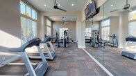 This single-floor fitness center is thoughtfully designed to support a comprehensive workout experience. The space is defined by soaring ceilings and an abundance of natural light streaming in through large, multi-pane windows positioned at both standard and clerestory heights, creating a bright and energizing atmosphere throughout the day. Warm terracotta-toned accent walls contrast with neutral gray tones to give the room a dynamic yet welcoming character.

The main workout floor is finished with durable, patterned carpet tiles in a multi-tone earthy palette, providing both comfort and practicality underfoot. Ceiling fans are strategically placed throughout to maintain airflow during intense workouts. Wall-mounted flat-screen televisions offer entertainment while exercising.

The cardio section is well-stocked with Precor treadmills and upright stationary bikes, all arranged to face the windows and mirrors for an open, spacious feel. A dedicated strength training area features a multi-station cable machine along with a free-weight rack and adjustable workout bench. Full-length mirrors line one wall, visually expanding the space and allowing users to monitor their form. A water dispenser and waste receptacle are conveniently located within the space for easy access, rounding out the practical amenities of this well-appointed fitness facility.