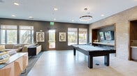 This contemporary game room delivers a dynamic, multi-functional entertainment experience across a generously sized single-floor layout. The space is anchored by a sleek pool table positioned at the center, complemented by a ping-pong table and a communal game table set with chairs — ideal for board games and casual gatherings. A built-in wet bar with cabinetry and a mini-fridge is seamlessly integrated into a textured light brick accent wall, framed by warm wood surrounds that add architectural depth and contrast. Wall-mounted flat-screen TVs are thoughtfully positioned throughout, including within a cozy nook featuring a striped built-in bench, perfect for spectators. Large-format tile flooring runs throughout the main activity area, while plush carpet defines the comfortable lounge zone, where a generous sectional sofa faces mounted displays — an inviting retreat within the broader space. Circular pendant light fixtures cast a warm, modern glow overhead, supplemented by recessed lighting. Bold abstract artwork and a large statement painting add personality to the walls. Expansive windows and a glass door flood the space with natural light and provide direct access to an outdoor patio, extending the entertainment area beyond the interior.
