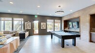 This contemporary game room delivers a dynamic, multi-functional entertainment experience across a generously sized single-floor layout. The space is anchored by a sleek pool table positioned at the center, complemented by a ping-pong table and a communal game table set with chairs — ideal for board games and casual gatherings. A built-in wet bar with cabinetry and a mini-fridge is seamlessly integrated into a textured light brick accent wall, framed by warm wood surrounds that add architectural depth and contrast. Wall-mounted flat-screen TVs are thoughtfully positioned throughout, including within a cozy nook featuring a striped built-in bench, perfect for spectators. Large-format tile flooring runs throughout the main activity area, while plush carpet defines the comfortable lounge zone, where a generous sectional sofa faces mounted displays — an inviting retreat within the broader space. Circular pendant light fixtures cast a warm, modern glow overhead, supplemented by recessed lighting. Bold abstract artwork and a large statement painting add personality to the walls. Expansive windows and a glass door flood the space with natural light and provide direct access to an outdoor patio, extending the entertainment area beyond the interior.