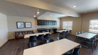 This single-floor multipurpose room is thoughtfully designed to accommodate a wide range of activities, from arts and crafts to group gatherings. The open layout features multiple large white rectangular tables paired with black stackable chairs, providing ample seating and workspace for participants. Dark hardwood-style flooring runs throughout, complementing the warm, neutral-toned walls and creating a cohesive, inviting atmosphere.

A standout feature of the room is the expansive wall of dark-stained open shelving units, which offer extensive storage for supplies, books, bins, and project materials, keeping the space organized and functional. Natural light filters in through multiple windows fitted with horizontal blinds, brightening the interior and offering views to the surrounding exterior.

Along one wall, a built-in kitchenette area includes dark espresso-stained upper cabinetry and lower drawers flanking a double-basin sink with a gray countertop — ideal for project cleanup or light refreshments. Framed artwork adorns the walls, adding a decorative touch to the otherwise utilitarian setting.

Recessed ceiling lighting provides consistent illumination throughout, while the varying ceiling heights, including a raised section, add architectural interest to the space. Overall, this room is well-suited for community programs, workshops, and creative activities.