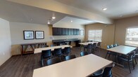 This single-floor multipurpose room is thoughtfully designed to accommodate a wide range of activities, from arts and crafts to group gatherings. The open layout features multiple large white rectangular tables paired with black stackable chairs, providing ample seating and workspace for participants. Dark hardwood-style flooring runs throughout, complementing the warm, neutral-toned walls and creating a cohesive, inviting atmosphere.

A standout feature of the room is the expansive wall of dark-stained open shelving units, which offer extensive storage for supplies, books, bins, and project materials, keeping the space organized and functional. Natural light filters in through multiple windows fitted with horizontal blinds, brightening the interior and offering views to the surrounding exterior.

Along one wall, a built-in kitchenette area includes dark espresso-stained upper cabinetry and lower drawers flanking a double-basin sink with a gray countertop — ideal for project cleanup or light refreshments. Framed artwork adorns the walls, adding a decorative touch to the otherwise utilitarian setting.

Recessed ceiling lighting provides consistent illumination throughout, while the varying ceiling heights, including a raised section, add architectural interest to the space. Overall, this room is well-suited for community programs, workshops, and creative activities.