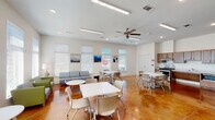 Located in Covington, Louisiana, this single-floor community room offers a welcoming, versatile gathering space designed for both casual socializing and organized events. The room is defined by its rich, stained concrete flooring, which adds warmth and visual depth while remaining highly durable and easy to maintain.

The main open area is flooded with natural light through multiple large windows fitted with blinds, creating a bright and airy atmosphere. The white walls are accented with colorful artwork, contributing a lively yet clean aesthetic. Recessed lighting and linear flush-mount fixtures supplement natural illumination, while a ceiling fan promotes comfortable airflow throughout the space.

Seating is arranged flexibly, combining multiple white rectangular tables with mobile chairs to accommodate various group sizes and activities. A lounge area along one wall features upholstered sofas and accent chairs in green and gray tones, providing a comfortable spot for informal gatherings.

A fully equipped kitchen area lines one wall, outfitted with warm wood cabinetry, a stainless steel range, built-in microwave, full-size refrigerator, and a sink with counter space—ideal for catering or community events. A set of glass double doors provides direct access to an outdoor area, further enhancing the sense of openness and connectivity within this functional community space.
