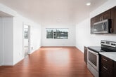 This single-floor unit offers a clean, modern aesthetic defined by crisp white walls, warm hardwood-style flooring, and an abundance of natural light throughout. The open-concept living and dining area is spacious and airy, anchored by a large window that frames views of the surrounding neighborhood and fills the space with natural light. The seamless flow between the living area and kitchen makes the layout feel expansive and functional.

The kitchen is well-equipped for everyday living, featuring rich dark wood cabinetry with ample upper and lower storage, light-colored countertops, a stainless steel sink, and a full suite of stainless steel appliances including an electric range, microwave, dishwasher, and refrigerator. Recessed lighting adds a polished touch to the cooking space.

The bathroom is neatly finished in a bright, all-white palette, complete with a full soaking tub and shower combination, a toilet, and a dark wood-framed vanity mirror that complements the cabinetry found throughout the unit. The bedroom features plush carpeting for comfort underfoot and includes a sliding-door closet with wire shelving for organized storage. Together, the rooms create a cohesive, comfortable living environment with a consistent modern design language.