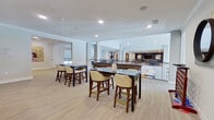 This two-story communal space is thoughtfully designed to offer a variety of gathering and recreational areas across both levels. The layout is anchored by a dramatic double-height atrium, where soaring ceilings and expansive windows flood the interior with natural light, creating an airy, welcoming atmosphere throughout.

The upper level features an open lounge and seating area with contemporary armchairs and side tables positioned along a railed mezzanine overlooking the floor below. High-top bar tables with upholstered seating provide casual gathering spots, while the open sightlines between floors enhance the sense of connectivity and spaciousness. Curated abstract artwork is displayed prominently along the walls, adding vibrant color and visual interest.

A dedicated games room on this level is equipped with a billiards table set against a deep navy accent wall, as well as a separate poker room featuring a round game table surrounded by plaid upholstered chairs and a wall-mounted television. An elevator provides convenient access between floors.

Light wood-tone flooring runs throughout, complementing the neutral palette of white walls and gray structural columns. Recessed lighting and crown molding add refined architectural detail, contributing to an overall ambiance that is both polished and inviting.