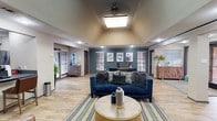 This well-appointed single-floor space functions as a community clubhouse or leasing office, blending modern design with warm, inviting touches throughout. The centerpiece of the main lounge is a soaring vaulted ceiling with a large skylight and ceiling fan, flooding the open area with natural light. Rich wood-look flooring runs throughout, complementing a bold navy blue sofa, a striped area rug, and a gold-toned circular coffee table that anchor the seating area. A striking stacked stone fireplace with a wall-mounted television serves as a dramatic focal point on one end of the lounge, flanked by leather armchairs for a cozy gathering space. The teal-painted accent wall at the rear of the lounge is adorned with framed watercolor artwork, adding a cohesive artistic flair to the space. A dedicated private office area features a substantial wood desk, velvet blue guest chairs, and large windows that allow ample natural light. Adjacent to the lounge, a functional kitchenette with a gray quartz countertop bar, leather bar stools, open shelving, and stainless steel appliances provides a convenient refreshment station. A restroom is also accessible from this area. Overall, the space exudes a polished, contemporary ambiance with a warm and welcoming character.