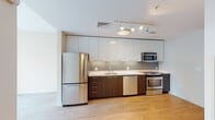 This well-appointed single-floor unit offers a clean, modern aesthetic carried consistently throughout the space. Light hardwood flooring runs continuously from the entry hallway through the living area, lending a cohesive and airy feel to the interior.

The entryway corridor is lined with closet storage, providing generous organization space before opening into the main living area. The living space is bathed in natural light, courtesy of a large floor-to-ceiling window that frames views of the surrounding urban cityscape, with vertical blinds offering privacy when needed. A ceiling fan provides additional comfort to this bright, open area.

The kitchen is a standout feature, designed in a contemporary galley style with a striking two-tone cabinet scheme — high-gloss white upper cabinets contrast elegantly with rich dark wood lower cabinetry. A grey subway tile backsplash adds texture, while white countertops provide a clean working surface. The kitchen is fully equipped with a full suite of stainless steel appliances, including a refrigerator, dishwasher, electric range, and built-in microwave, complemented by track lighting overhead.

The bathroom is finished in a neutral, warm palette with light hardwood-style flooring. It features a full soaker tub and shower combination with built-in shelving, a single vanity with a dark wood base, and chrome fixtures and grab bars for added accessibility.