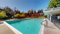 Located at 400 E Remington Dr in Sunnyvale, California, this outdoor pool area serves as a central communal amenity within a well-established residential community in the heart of Silicon Valley. The centerpiece is a generously sized swimming pool with clear turquoise water, framed by a wide concrete pool deck that provides ample space for lounging and recreation. Sun chairs and lounge seating are arranged along the poolside, offering comfortable spots to relax under the abundant California sunshine. A covered walkway runs along one side of the pool area, supported by sturdy columns and providing shaded access along the perimeter. The space is enclosed by a secure metal fence, ensuring safety and privacy for residents. Vibrant red umbrellas accent an outdoor seating and dining area adjacent to the pool, creating a welcoming social space. Mature trees — including large leafy specimens and tall conifers — surround the area, offering natural shade and a lush, park-like atmosphere. The multi-story residential buildings visible in the background feature warm-toned exterior finishes with balconies, complementing the inviting outdoor environment. The overall ambiance is bright, open, and resort-like, making this pool area a standout feature of the community.