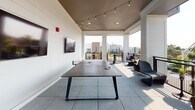 Located at 1425 Semmes Ave in Richmond, Virginia's Manchester neighborhood, this single-level covered outdoor lounge offers a well-appointed communal retreat with open city views. The space features large-format light gray tile flooring and a dark ceiling accented by warm Edison-style string lights, creating an inviting ambiance suitable for both daytime and evening use.

A centerpiece ping-pong table anchors the interior portion of the lounge, accompanied by two wall-mounted flat-screen TVs displaying local Richmond imagery. The shiplap-clad white walls lend a clean, modern aesthetic throughout. Open-air sections extend toward the perimeter railing, where yellow bar-height tables and chairs provide bright pops of color, while wicker lounge chairs and side tables offer a more relaxed seating option.

The space transitions seamlessly between its covered interior and open-air terrace through wide column-framed openings, allowing abundant natural light and panoramic views of the surrounding Richmond skyline and neighboring developments. A teal-framed entry door adds a distinctive architectural detail. Overall, the lounge balances a casual, recreational atmosphere with thoughtful design elements suited to social gatherings.