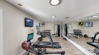 This dedicated home gym is thoughtfully designed to deliver a full fitness center experience within a private setting. The room features light wood-look flooring throughout, white walls, and a textured ceiling with flush-mounted LED lighting that keeps the space well-lit and functional. A large wall-to-wall mirror runs along one side of the room, visually expanding the space and allowing users to monitor their form during workouts.

The cardio zone is well-represented, with Echelon treadmills, a rowing machine, and smart spin bikes, each equipped with interactive touch screens for guided workout sessions. A wall-mounted flat-screen TV and a wall fan add to the comfort and entertainment options during training.

The strength training area includes a Hoist cable machine system, an adjustable weight bench, a full rack of hex dumbbells, and a barbell with plates, providing ample options for resistance and free weight training. French doors connect the gym to an adjacent living area, and a window allows natural light to filter in.

Overall, the space is organized efficiently, balancing cardio and strength equipment without feeling overcrowded, making it a comprehensive and practical private fitness facility.