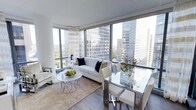 This single-floor high-rise residence offers a refined, open-concept layout that seamlessly connects the living and dining areas. The space is defined by expansive floor-to-ceiling windows that wrap around the exterior walls, flooding the interior with abundant natural light and framing panoramic views of the surrounding urban skyline, including neighboring high-rise towers and patches of green treetops below.

The living area is anchored by a plush, cream-colored sofa accented with navy blue throw pillows, complemented by a sleek, round marble-top coffee table with a metallic base. A neutral toned area rug with subtle detailing grounds the seating arrangement, adding warmth to the light hardwood flooring that runs throughout.

Adjacent to the living space, the dining area features a glass-top table with a polished chrome frame, paired with elegant upholstered white dining chairs. Sheer striped curtains frame the windows, softening the abundant natural light without obstructing the impressive city views. The overall ambiance is polished and contemporary, with a cohesive neutral palette punctuated by metallic accents that lend a sophisticated, cosmopolitan character to the space.