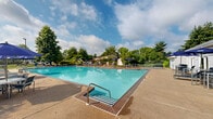 Located at 9071 Mill Creek Rd in Levittown, Pennsylvania, this outdoor recreation facility centers around a generously sized swimming pool with crystal-clear turquoise water. The pool is framed by a wide concrete sundeck furnished with lounge chairs and blue patio umbrellas, providing ample space for relaxation and socializing. Entry to the pool is made easy via stainless steel handrails and a step-in entry point, accommodating swimmers of all abilities.

The adjacent single-story clubhouse is clad in white lap siding and features a gabled roof with skylights. A covered patio extends from the building directly toward the pool deck, outfitted with additional seating and umbrella-shaded tables — ideal for outdoor gatherings. Large French doors connect the interior of the clubhouse to the pool area, facilitating a seamless indoor-outdoor flow.

The entire pool area is enclosed by a safety fence, and the surrounding landscape is lush with mature deciduous and evergreen trees that provide natural shade and privacy. Additional amenities visible on the grounds include what appears to be a tennis or sport court and a playground structure, rounding out this well-equipped community recreation space. The open sky and verdant surroundings contribute to an inviting and refreshing outdoor atmosphere.