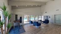 This single-floor community clubhouse or leasing center is defined by its striking architectural character, most notably the soaring vaulted ceilings with exposed white-painted beams and wood plank detailing that lend the space a bright, airy feel. Track lighting runs along the ceiling beams, illuminating the open-plan interior with a clean, modern glow complemented by abundant natural light streaming through expansive floor-to-ceiling windows and glass entry doors. The main reception area features a sleek, contemporary front desk set against a bold blue accent wall adorned with framed artwork and a mounted flat-screen television. A beverage and amenity bar lines the back wall, creating a welcoming hospitality touch. The lounge area is furnished with navy blue armchairs arranged around low tables atop patterned area rugs, offering a comfortable gathering space. Large picture windows frame direct views of a shimmering outdoor swimming pool and mature trees, seamlessly connecting the interior to the outdoor environment. Wide-plank wood-look flooring flows throughout the entire space, unifying the various functional zones. Additional private rooms, including what appears to be a small cinema or media room and a private office, are accessible off the main floor, providing versatile functionality within this well-appointed community facility.