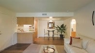 This single-floor residence showcases an open-concept layout that seamlessly connects the living, dining, and kitchen areas into one cohesive and functional space. The living area is anchored by a plush white sofa paired with a decorative coffee table, creating a comfortable and inviting atmosphere. Warm hardwood-style flooring flows through the central and dining areas, while a large area rug defines the living space.

The dining area features a compact round bistro-style table with wrought iron chairs, positioned beneath a pendant light fixture that draws the eye upward to a subtly tray-accented ceiling. A decorative framed mirror and a lush indoor tropical plant add character and a natural element to the space.

The kitchen is partially open to the main living area, separated by a raised countertop breakfast bar. Light wood cabinetry lines the upper and lower sections of the kitchen, complementing the white appliances visible in the background. An archway on the right side of the space hints at additional rooms beyond. The textured ceiling and neutral wall tones contribute to a clean, bright, and well-organized overall ambiance.
