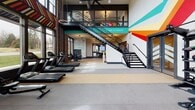 Located on Otterdale Road in Chesterfield, Virginia, this two-story fitness facility delivers a dynamic, energizing environment designed to motivate at every turn. The ground floor opens into a expansive main workout area defined by soaring double-height ceilings, exposed brick walls, and large windows that flood the space with natural light and offer open views of the surrounding landscape. Bold, colorful geometric murals and motivational typography cover the walls, creating a visually stimulating atmosphere throughout. The main floor is divided into distinct training zones: a cardio section outfitted with treadmills, rowing machines, stationary bikes, and ellipticals; a functional training area featuring kettlebells, battle ropes, medicine balls, and plyo boxes on durable rubber flooring; and a strength zone with selectorized machines and benches. A striking open staircase with black metal railings connects the two levels, its risers emblazoned with motivational phrases. The upper level offers a studio space with wide-plank hardwood flooring, floor-to-ceiling mirrors, cycling bikes, and suspended circular pendant lighting alongside exposed ductwork, lending an industrial-chic aesthetic. A mezzanine overlook connects back to the main floor below, reinforcing the facility's sense of open, connected space.