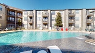 This outdoor amenity space is part of a contemporary multi-story residential community designed with both aesthetics and functionality in mind. The architecture features a neutral palette of beige and gray tones, horizontal lap siding, and large windows, giving the building a clean, modern appearance with private balconies overlooking the shared grounds below.

The centerpiece of the outdoor space is a generously sized resort-style swimming pool with clear blue water, surrounded by a wide concrete deck furnished with lounge chairs. Bold red decorative planters add striking color accents throughout the courtyard, complementing the lush landscaping that includes mature trees and low ground cover plantings.

A dedicated pet area is enclosed by sturdy dark metal fencing with a mesh panel design, featuring artificial turf for year-round use. The enclosure offers a secure and practical space for residents with pets. Nearby, concrete steps with natural stone retaining walls and dark metal handrails provide a well-designed transition between different levels of the property.

The common corridors and transition spaces feature concrete masonry walls, large grid-patterned windows, and gated access points, all contributing to a cohesive and secure environment. String lights accent the outdoor areas, enhancing the ambiance during evening hours. Overall, the space balances practicality with inviting, resort-inspired outdoor living.