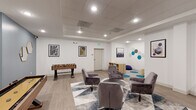 Located at 211 Warren Ave in Baltimore, MD, this single-floor amenity space is thoughtfully designed to serve as a multifunctional hub for recreation, socializing, and productivity. The interior features light hardwood-style flooring throughout, recessed lighting with a tray ceiling detail, and crisp white walls that create a bright, open atmosphere.

The space is divided into distinct functional zones. A central lounge area anchors the room with a cluster of plush grey swivel chairs arranged around a sleek round coffee table, all set atop a geometric blue and grey area rug that adds visual interest underfoot. Adjacent to the lounge, a gaming zone offers both a foosball table and a shuffleboard table, providing ample entertainment options. A dedicated children's or activity corner features open wood cubby shelving and playful teal hexagonal tile flooring accents that echo the bold hexagonal wall art displayed prominently throughout the space.

On the opposite end, a conference and collaborative work area is equipped with a long communal table lined with modern blue bar-height chairs, positioned beneath a wall-mounted flat-screen television — ideal for meetings or presentations. Framed artwork adds personality to the walls, complementing the space's contemporary aesthetic. The overall design strikes a balance between energetic and inviting, making it a versatile destination for both leisure and work.