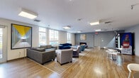 This single-floor communal lounge offers a generously sized, open-plan layout designed to accommodate a variety of social and leisure activities. The space is finished with warm wood-look flooring throughout and illuminated by a series of flush-mounted square panel lights that provide consistent, even lighting across the entire area.

The main lounge zone features multiple distinct seating arrangements, including a large sectional sofa, a navy blue couch, and an assortment of upholstered armchairs grouped around low tables — ideal for both casual conversation and relaxed gathering. A sleek, wall-mounted electric fireplace anchors one end of the lounge, framed by a tiled surround and complemented by a colorful abstract artwork above it. Large windows along one wall admit abundant natural light and offer views of the surrounding exterior.

Adjacent to the main lounge is a kitchenette area, outfitted with dark cabinetry, granite-style countertops, a stainless steel refrigerator, and a sink — providing practical utility for residents or guests. A vibrant yellow accent wall and framed artwork give this area a distinct, cheerful character.

A dedicated vending area near the exit features snack and beverage dispensers, adding everyday convenience to the space. The overall ambiance is modern and welcoming, with contemporary furnishings and curated artwork contributing to a clean, community-focused aesthetic.