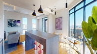 Located at 337 Seventh Street West in Saint Paul, Minnesota, this contemporary communal space offers a thoughtfully designed, open-plan layout spread across a single level. The living area is anchored by a sleek linear fireplace set into a dark, textured accent wall, complemented by a wall-mounted television and ambient cove lighting recessed into a tray ceiling above. Warm hardwood flooring flows throughout, unifying the space and enhancing its inviting atmosphere.

Flexible seating arrangements — including a leather sofa, modular hexagonal ottomans, and accent chairs — encourage a variety of uses, from casual lounging to social gatherings. A large concrete-topped bar-height island serves as the centerpiece of the social area, surrounded by modern counter stools and pendant lighting in bold black finishes. Large floor-to-ceiling windows on multiple sides flood the interior with natural light and frame striking views of the Saint Paul skyline, including a notable view of a historic domed building.

Curated contemporary artwork adorns the walls, adding creative character throughout. A standout feature is the adjoining rooftop terrace, accessible via full-height glass doors, featuring outdoor seating, a striking graphic mural, and panoramic views of the surrounding cityscape and natural landscape beyond.