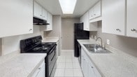 This single-floor residential unit offers a straightforward and functional layout designed for comfortable everyday living. The open living and dining area transitions seamlessly between light tile flooring near the entry and soft wall-to-wall carpeting, creating a welcoming atmosphere. Neutral gray walls and white baseboards are consistent throughout, lending the space a clean, cohesive look. A window with blinds allows natural light to filter into the living area.

The galley-style kitchen is well-equipped, featuring black appliances including an electric range and refrigerator, a stainless steel sink, light granite-style countertops, and ample white cabinetry above and below. A skylight-style overhead fixture brightens the kitchen corridor. The kitchen connects to a utility area at the rear.

The bedrooms are carpeted and finished in the same neutral gray palette, each with a window fitted with blinds for natural light and privacy. The rooms offer comfortable proportions suitable for standard bedroom furnishings.

The bathroom includes a full tub and shower combination with white tile surround, chrome fixtures, and a clean, minimalist finish. Overall, the unit presents a well-maintained, move-in-ready space with a consistent design aesthetic throughout.