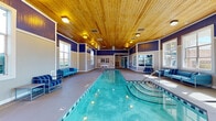 Located at 200 Edinborough Dr in Durham, NC, this single-story indoor aquatic facility offers a well-appointed swimming environment designed for both recreation and relaxation. The centerpiece is a generous lap pool with clearly marked lanes, complemented by a separate spa with stepped entry, both finished in vibrant aqua tones. The space is crowned by a warm, natural wood-planked ceiling that lends a distinctive architectural character, contrasting elegantly with the bold navy blue upper walls and crisp white wainscoting below. Abundant natural light pours in through large, multi-pane windows positioned around the perimeter, offering views of the surrounding landscaped grounds and creating an open, airy atmosphere throughout. Scattered around the pool deck are modern metal-framed seating arrangements upholstered in blue-striped and solid blue cushions, providing comfortable lounging areas for guests. A dedicated observation and lounge area with floor-to-ceiling windows overlooks both the indoor pool and an outdoor pool area, making it a versatile social space. Safety signage, a wall-mounted life ring, and poolside equipment are thoughtfully integrated throughout. The smooth concrete pool deck and well-maintained finishes reflect a commitment to both functionality and comfort in this inviting aquatic retreat.