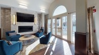 Located at 800 Central in Salem, New Hampshire, this beautifully designed clubhouse serves as a sophisticated social hub for the community. The space is defined by a striking mix of modern and warm design elements throughout its open, single-level layout.

The main lounge area commands attention with soaring vaulted ceilings and a dramatic arched window that floods the room with natural light while offering views of the surrounding community. A sleek, stone-clad linear fireplace anchors the seating area, complemented by plush blue accent chairs, patterned rugs, and decorative horseshoe-motif wall art that nods to local character. Wood-look tile flooring runs throughout, adding warmth and continuity to the space.

A well-appointed wet bar area features rich dark wood cabinetry, open shelving with under-cabinet lighting, a textured dimensional tile backsplash, a mounted flat-screen TV, and a generous bar counter with yellow upholstered seating — ideal for casual gatherings.

The game room offers a dedicated shuffleboard table and wall-mounted television, providing an inviting space for recreation. Adjacent lounge nooks with built-in shelving and curated artwork create quieter retreat areas for residents.

Wide, light-filled corridors with clean white walls and warm wood-accented columns connect the various areas seamlessly, creating a cohesive and welcoming environment throughout.