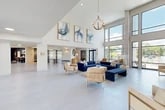 Located at 4781 SW 36 Rd in Gainesville, FL, this single-floor community clubhouse offers a generously sized, open-concept layout designed to impress and accommodate. The space is anchored by dramatic double-height ceilings that rise in multiple areas, most notably in a striking rotunda-style lounge featuring a bold navy-painted ceiling with pendant lighting and floor-to-ceiling windows that flood the interior with natural light and offer views of the surrounding landscape.

Throughout the space, large-format light gray tile flooring provides a clean, contemporary foundation. The main lounge area is elegantly appointed with deep navy blue velvet sectional sofas, neutral accent chairs, and a stone-topped coffee table, all complemented by a curated selection of abstract framed artwork. A textured feature wall adds warmth and visual depth near the entryway, where a mezzanine-level railing overlooks the ground floor below.

Toward the rear, a fully equipped kitchenette with cabinetry, a sink, and appliances serves residents, while an adjacent business center offers bar-height workstations with computer monitors set against a richly toned accent wall. An orb-style chandelier and woven pendant lights add refined character throughout. Large glass entry doors and expansive windows ensure seamless indoor-outdoor connectivity throughout this well-appointed amenity space.