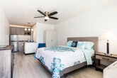 This single-floor studio offers a thoughtfully arranged open-concept layout that efficiently combines living, sleeping, and cooking areas into one cohesive space. The main living area is anchored by a comfortable bed, complemented by a ceiling fan with a built-in light fixture that helps keep the space airy and well-ventilated. Light wood-style flooring runs throughout, contributing to the bright and clean aesthetic reinforced by white walls and neutral tones.

The kitchen is a standout feature, outfitted with gray shaker-style cabinetry, a stainless steel refrigerator, a built-in microwave, and a sink. A peninsula with a granite-style countertop provides additional workspace and doubles as a casual dining area, illuminated by a stylish pendant light fixture overhead. A freestanding white wardrobe offers convenient storage within the sleeping area, while a rustic wood dresser adds warmth and character to the space.

The bathroom is clean and functional, finished in crisp white tones with light flooring, a toilet, and a towel bar, maintaining the consistent, refreshed aesthetic found throughout the unit. Overall, this studio presents a compact yet complete living environment with a cohesive coastal-inspired color palette of whites, blues, and warm wood accents.
