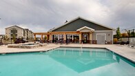 Located in Spanish Fork, Utah, this outdoor amenity area offers a resort-style retreat designed for relaxation and recreation. The centerpiece is a generously sized swimming pool with crystal-clear water, complemented by a separate raised hot tub spa, providing options for both active swimming and leisurely soaking. The surrounding concrete sundeck is expansive, lined with rows of lounge chairs and accented by wicker seating sets with slatted tables, offering ample space for sunbathing and outdoor socializing. A charming pergola-covered structure anchors one side of the pool area, featuring large glass panel doors that open to an interior clubhouse space — its board-and-batten siding and brick accents giving it a contemporary yet welcoming architectural character. The entire area is enclosed by a combination of metal fencing and privacy panels, creating a secure and intimate atmosphere. Tall light poles ensure the space is well-lit for evening use. Sweeping views of the surrounding Utah mountain ranges are visible beyond the perimeter, adding a dramatic natural backdrop to this well-appointed outdoor community amenity.