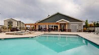 Located in Spanish Fork, Utah, this outdoor amenity area offers a resort-style retreat designed for relaxation and recreation. The centerpiece is a generously sized swimming pool with crystal-clear water, complemented by a separate raised hot tub spa, providing options for both active swimming and leisurely soaking. The surrounding concrete sundeck is expansive, lined with rows of lounge chairs and accented by wicker seating sets with slatted tables, offering ample space for sunbathing and outdoor socializing. A charming pergola-covered structure anchors one side of the pool area, featuring large glass panel doors that open to an interior clubhouse space — its board-and-batten siding and brick accents giving it a contemporary yet welcoming architectural character. The entire area is enclosed by a combination of metal fencing and privacy panels, creating a secure and intimate atmosphere. Tall light poles ensure the space is well-lit for evening use. Sweeping views of the surrounding Utah mountain ranges are visible beyond the perimeter, adding a dramatic natural backdrop to this well-appointed outdoor community amenity.