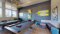 This well-appointed game room is designed to serve as a dynamic social and recreational hub. The space accommodates two full-sized pool tables with slate-gray felt surfaces and dark wood frames, complemented by a shuffleboard table positioned along one wall beneath a wall-mounted flat-screen television. A cue rack is conveniently mounted nearby for easy access.

The flooring is a standout design element, combining wood-look planks with an eye-catching geometric hexagonal tile inlay, adding visual depth and texture throughout. One accent wall is covered in a bold wave-patterned wallcovering in a cool blue-gray tone, serving as a backdrop for a curated collection of vibrant, colorful abstract paintings that energize the atmosphere.

Soaring ceilings with large-format windows along one wall allow generous natural light to flood the space, offering outdoor views and a sense of openness. A ceiling fan overhead ensures comfortable airflow. Seating areas include a sectional sofa, rounded ottomans in blue, and a pair of sleek swivel chairs arranged around a drum-style side table, offering comfortable spots to relax between games. The overall ambiance is modern, lively, and inviting.