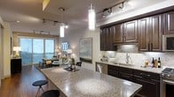 This single-floor residence showcases a thoughtfully designed open-concept layout that seamlessly connects the kitchen, dining, and living areas. The kitchen is a standout feature, outfitted with rich, dark espresso cabinetry, light granite countertops, a decorative tile backsplash, and stainless steel appliances including a dishwasher. A generous kitchen island with bar seating provides an ideal space for casual dining and entertaining. Pendant lighting with sleek cylindrical glass fixtures hangs above the island, while an adjustable track lighting system illuminates the entire open living space. The dining area flows naturally from the kitchen, featuring an elegant table setting with fresh floral accents. The living area beyond benefits from large floor-to-ceiling windows that invite abundant natural light and frame a picturesque outdoor view of the surrounding landscape and waterway. Neutral wall tones and warm wood flooring throughout create a cohesive and inviting ambiance. The overall design balances modern sophistication with comfortable livability, making it an ideal space for both everyday living and entertaining guests.