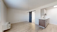 Located at 2131 S Swope Dr in Independence, MO, this thoughtfully updated one-bedroom, one-bathroom unit makes smart use of its compact footprint across a single floor. Light-toned vinyl plank flooring runs throughout the main living areas, contributing to a bright and cohesive aesthetic.

The open living area is finished with crisp white walls and a textured ceiling, and features a wall-mounted PTAC unit for climate control. A standout sliding barn door with a sleek black hardware track serves as a stylish divider, connecting the living space to the rest of the unit. Natural light filters in through a window fitted with blinds.

The galley-style kitchen is efficiently designed, offering ample white cabinetry with modern hardware, quartz-look countertops, a stainless steel sink with a matte black faucet, and black appliances including an electric range and refrigerator.

The bedroom is carpeted for comfort and includes a ceiling fan with a light fixture, a built-in closet with wire shelving, and direct access to the bathroom. The bathroom features a white vanity with a stone-look countertop, an integrated sink, a large mirror with modern lighting, and matte black hardware accents throughout, maintaining a consistent modern design language across the entire unit.