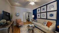 Located at 1100 Legends Club Dr in the desirable community of Mt. Pleasant, South Carolina, this single-floor home offers a thoughtfully designed interior that blends comfort with refined style. The living area serves as a centerpiece of the home, showcasing a striking navy blue accent wall adorned with a gallery of framed botanical artwork in gold-toned frames. Warm hardwood flooring flows throughout, complemented by neutral walls trimmed with crisp crown molding. A ceiling fan overhead ensures year-round comfort, while an elegant arched doorway leads seamlessly into adjacent spaces, adding architectural character to the layout. The living room is furnished with a plush neutral sofa accented with patterned throw pillows, a vibrant floral accent chair, and a distinctive glass-top coffee table with a natural wood base, all anchored by a layered area rug. A decorative sunburst mirror and carefully placed accent lighting contribute to the warm, curated ambiance. The arched entryway glimpsed in the background hints at additional well-appointed spaces beyond, reinforcing the cohesive and polished aesthetic carried throughout this single-level home.