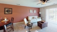 This single-story residence located on N Brickyard Road in Columbia, South Carolina, offers a well-organized layout with distinct living spaces connected by carpeted hallways. The living room serves as the heart of the home, distinguished by a warm terracotta accent wall, plush carpet flooring, and a ceiling fan with light fixture. The space flows naturally into a dedicated dining area, where a pendant light anchors a raised dining table set, complemented by the same accent wall color and an open view into the kitchen. The kitchen features warm wood cabinetry extending to the ceiling, generous counter space, a coil-element range, and light hardwood-style flooring, providing both functionality and storage capacity. A pass-through connects the kitchen to the dining area, enhancing the open feel. The bedroom is bright and carpeted, with a window admitting natural light, a closet, and space for a dedicated desk workstation. Two full bathrooms each include tile flooring, a single-sink vanity with wood cabinetry, a large mirror, and a tub-shower combination. A dedicated laundry room houses a full-sized washer and dryer with an overhead shelf, adding convenient in-unit utility.