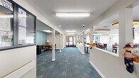 Located at 1001 Leland in Detroit, Michigan, this single-floor community facility offers a generously sized, well-appointed space designed to support group activities and daily living. The layout is anchored by a broad, open common area that serves as the heart of the facility. Distinctive wood-framed overhead light fixtures illuminate the vaulted ceiling, while large windows along the perimeter flood the room with natural light and contribute to its bright, welcoming ambiance. The floors feature a combination of patterned blue-toned carpeting and smooth hardwood, adding visual warmth throughout. Multiple seating arrangements with tables and chairs accommodate group gatherings, dining, and programming activities. Teal accent walls provide a calming contrast to the predominantly white interior. A staffed reception counter is integrated into the main area, allowing for easy oversight and resident interaction. The wide, column-lined hallway connects the entrance to the various functional areas, including a dedicated exercise room and a laundry facility equipped with a utility sink and cabinetry. The entrance provides a clear and accessible point of entry. Overall, the space is thoughtfully organized to balance community engagement with practical, everyday functionality.