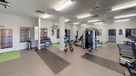 This single-floor fitness center is thoughtfully designed to accommodate a wide range of exercise disciplines. The space is organized into distinct functional zones, each catering to different training needs. The main workout area features bright, fluorescent overhead lighting, ceiling fans, and light-toned walls accented by a teal blue feature wall, creating an energizing yet comfortable atmosphere.

The cardio zone houses multiple treadmills and an elliptical trainer, positioned near large windows that allow natural light to fill the space. Adjacent to this area, a dedicated strength training section includes a cable machine, power rack with weight plates, and a multi-station functional trainer, all set on protective interlocking foam and rubber flooring.

A separate functional training zone offers a TKO heavy punching bag, TRX suspension straps, battle ropes, medicine balls in various weights, kettlebells, a stability ball, and open mat space for floor exercises. Interlocking foam tiles in green and gray provide cushioning throughout this area.

A standout feature of the facility is the private golf simulator room, equipped with a SwingU projection screen and a turf hitting mat, accompanied by a seating area with high-top table and stools. An accessible restroom is also available on-site.