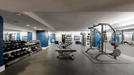 Located at 1211 W Ninth St in Cleveland's vibrant Ohio City neighborhood, this dedicated fitness center offers a thoughtfully designed exercise space across a single floor. The room features a striking aesthetic defined by bold blue-painted brick columns that contrast with clean white walls, creating a dynamic and energizing atmosphere. Durable rubber flooring covers the entire workout area, providing safety and comfort underfoot.

The space is comprehensively outfitted for a wide variety of training styles. A full dumbbell rack holds an extensive range of free weights, complemented by multiple adjustable benches for strength training. Overhead racks display kettlebells and medicine balls in various sizes, supporting functional and dynamic workouts. True-brand cable machines offer versatile resistance training options, while squat racks and barbell stations accommodate serious lifters.

Resistance bands and suspension training equipment are neatly organized and wall-mounted for convenient access. Large mirrors along the walls enhance the sense of space and allow users to monitor their form. Recessed lighting and perimeter accent lighting provide bright, even illumination throughout. A wall-mounted flat-screen television adds an entertainment element to the workout environment. The layout is efficiently organized to maximize usable floor space, making this fitness center a fully functional and professionally appointed facility.