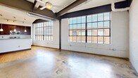 Unit 601 is a loft-style urban residence in Winston-Salem, NC, showcasing striking industrial character throughout. The open living area features polished concrete floors, exposed steel beams, white-painted brick walls, and large factory-style windows that flood the space with natural light and offer city views. A ceiling fan adds comfort to the airy layout. The kitchen is well-appointed with rich dark wood cabinetry, granite countertops, stainless steel appliances, a breakfast bar, and track lighting. Hardwood floors extend through the kitchen and hallways. The bathroom includes a granite vanity, tiled floors, and a tub/shower combo. Dual closets provide generous storage, and a dedicated laundry room with stacked washer/dryer adds everyday convenience.