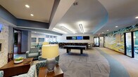 This single-story community amenity facility offers a diverse range of thoughtfully designed spaces. The centerpiece is a spacious game and social lounge featuring a pool table, a long bar-height counter with seating and multiple wall-mounted televisions, and a comfortable lounge area with patterned upholstered seating. A large-scale abstract mural adds a bold artistic focal point, while patterned carpet and recessed tray ceilings with linear lighting lend architectural interest throughout. Natural light enters through windows overlooking the surrounding community.

The fully equipped kitchen includes dark espresso cabinetry, granite countertops, a stainless steel dishwasher, and a undermount sink — well-suited for catering or events. The fitness center is generously sized and outfitted with a comprehensive array of cardio and strength equipment, accented by vibrant teal walls and large windows offering outdoor views.

A dedicated conference room provides a long central table, teal accent walls, and natural light from a row of windows, making it suitable for meetings or study sessions. Additional amenities include an accessible restroom with decorative stacked tile and an on-site laundry room with rows of washers and dryers on tile flooring.