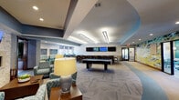 This single-story community amenity facility offers a diverse range of thoughtfully designed spaces. The centerpiece is a spacious game and social lounge featuring a pool table, a long bar-height counter with seating and multiple wall-mounted televisions, and a comfortable lounge area with patterned upholstered seating. A large-scale abstract mural adds a bold artistic focal point, while patterned carpet and recessed tray ceilings with linear lighting lend architectural interest throughout. Natural light enters through windows overlooking the surrounding community.

The fully equipped kitchen includes dark espresso cabinetry, granite countertops, a stainless steel dishwasher, and a undermount sink — well-suited for catering or events. The fitness center is generously sized and outfitted with a comprehensive array of cardio and strength equipment, accented by vibrant teal walls and large windows offering outdoor views.

A dedicated conference room provides a long central table, teal accent walls, and natural light from a row of windows, making it suitable for meetings or study sessions. Additional amenities include an accessible restroom with decorative stacked tile and an on-site laundry room with rows of washers and dryers on tile flooring.