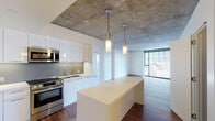 Located at the iconic 8500 W Sunset Blvd on the Sunset Strip in West Hollywood, this contemporary residence showcases a striking blend of industrial and modern design elements. The exposed concrete ceiling is a defining architectural feature throughout, lending an urban loft aesthetic that is complemented by warm, rich hardwood flooring underfoot.

The open-concept living and kitchen area serves as the heart of the space. The kitchen is outfitted with sleek white cabinetry, stainless steel appliances including a range and microwave, a subway tile backsplash, and a generous island with a clean white countertop. Cylindrical pendant lights hang above the island, providing focused illumination. The living area flows naturally from the kitchen, benefiting from large floor-to-ceiling windows that invite abundant natural light and offer views of the surrounding West Hollywood cityscape.

The bedroom is a serene retreat with the same hardwood flooring and exposed concrete ceiling, accompanied by a sliding glass door that opens to an exterior view. A dedicated hallway leads to a walk-in closet with built-in shelving, providing ample storage. The bathroom features a floating dark wood vanity with a white countertop and undermount sink, a backlit mirror, and clean white tile flooring, completing the space with a polished, minimalist finish.
