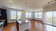 This single-floor unit is designed with a modern, open-concept layout that seamlessly connects the kitchen and living areas. The kitchen serves as the centerpiece, featuring dark espresso cabinetry that extends to the ceiling, a mosaic tile backsplash, stainless steel appliances including a refrigerator, range, and microwave, and a generous island with a granite countertop and undermount sink. Three distinctive yellow pendant lights hang above the island, adding a bold accent. Track lighting further illuminates the workspace.

The adjoining living area is bright and spacious, with warm hardwood-style flooring, a ceiling fan, and multiple windows fitted with white blinds that invite generous natural light and outdoor views. A hallway with panel doors connects the main living space to the private areas of the unit.

The bedrooms feature plush neutral carpeting, ceiling fans, and windows that allow natural light to filter in. One larger bedroom includes hardwood-style flooring and a sliding door, suggesting access to an outdoor area or closet.

The bathroom is well-appointed with dark cabinetry, granite countertops, and a large vanity mirror, while an adjacent walk-in closet provides wire shelving for ample storage. The overall design balances contemporary finishes with a comfortable, functional layout.