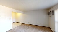 This single-floor unit offers a straightforward, functional layout connecting several distinct living areas. The spacious main living area is finished with neutral wall-to-wall carpeting and bright white walls, creating a clean, open atmosphere. Large sliding glass doors fitted with vertical blinds allow natural light to filter in, and a wall-mounted air conditioning unit provides climate control. Baseboard heating runs along the perimeter, ensuring efficient warmth throughout. A secondary carpeted room, accessible from the living area, similarly features white walls, vertical blinds, and ample natural light from a large window — suitable for use as a bedroom or additional living space. The galley-style kitchen is efficiently designed, featuring warm wood cabinetry on both sides, tile backsplash, light countertops with a stainless steel sink, and tile flooring. White appliances, including a refrigerator and range, are included. A door at the end of the kitchen provides additional access. The bathroom includes a full-size tub and shower combination with a white surround. Overall, the unit presents a clean, practical environment with a neutral palette well-suited for personalization.