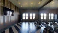 This single-floor fitness space combines bold design with practical functionality. The room is defined by its eye-catching chevron-patterned hardwood flooring, alternating between dark and light tones to create a dynamic visual effect that anchors the entire space. Dark-painted walls lend a dramatic, focused atmosphere well-suited to an exercise environment, while a large wall-mounted mirror expands the sense of space and allows users to monitor their form during workouts. Recessed ceiling lights are arranged in a uniform grid pattern, providing consistent, evenly distributed illumination throughout. Multiple windows along one wall invite natural light, brightening the interior and offering outside views. A wall-mounted flat-screen television adds a multimedia element for entertainment or guided workout sessions. The room is equipped with stationary cycling bikes and additional fitness equipment, making it a well-appointed exercise destination. The combination of refined architectural details—including the chevron floor, dark tones, and mirror—creates a fitness space that is both highly functional and visually sophisticated.