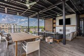 Located at 1399 Delashmut Ave in Columbus, Ohio, this contemporary rooftop amenity space seamlessly blends indoor comfort with outdoor living. The interior lounge is immediately distinguished by its bold orange ceiling, wood-look flooring, and large garage-style glass doors that retract to fully open the space onto the terrace. A wall-mounted TV, a woven hanging chair, and curated artwork contribute to the relaxed yet stylish atmosphere. A fully equipped kitchenette anchors one end of the interior, featuring dark wood cabinetry, a striking granite-topped island with bar seating, pendant lighting, and a stainless steel refrigerator — ideal for entertaining. The expansive covered terrace extends the living area outdoors, sheltered beneath an industrial-style steel pergola with ceiling fans and integrated lighting. Multiple seating arrangements, including lounge sofas and a long communal dining table, are arranged to take full advantage of sweeping views of the Columbus skyline and surrounding development. A dedicated outdoor kitchen zone, clad in warm wood paneling, provides a built-in grill and bar-height counter seating, perfect for al fresco gatherings. Large-format tile flooring unifies both the covered and open-air sections of the terrace, lending the entire space a cohesive, polished aesthetic.