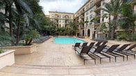 This expansive outdoor pool courtyard offers a resort-like retreat set within a beautifully designed Mediterranean-inspired complex. The centerpiece is a generously sized swimming pool with clear turquoise water, complemented by a separate hot tub nearby. Surrounding the pool, a wide stamped concrete deck provides ample space for relaxation, lined with rows of sleek black chaise lounges positioned to maximize comfort and sun exposure. At one end of the courtyard, private cabanas with rich burgundy curtains and canopy covers offer shaded, semi-enclosed retreats, each furnished with lounge chairs for a more intimate experience. The courtyard is lushly landscaped with tall palm trees, tropical plants, and dense greenery, creating a verdant atmosphere that enhances the sense of privacy and seclusion. The space is enclosed on multiple sides by a stately multi-story building featuring warm sandy-toned facades, arched windows, and private balconies that overlook the pool area. Decorative wall-mounted lanterns add a classic touch to the cabana structures. The overall ambiance is one of upscale leisure, seamlessly blending architectural elegance with tropical resort-style amenities in a well-maintained, cohesive outdoor environment.