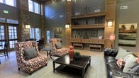 This single-story space makes a strong architectural impression with its dramatically high ceilings that amplify the sense of openness and grandeur. A striking stone accent column anchors one corner of the room, adding a natural, textural element that complements the warm, wood-toned built-in shelving unit along the back wall. The shelving spans floor to ceiling and features multiple display levels, drawers, and decorative ironwork accents, serving as a commanding focal point of the space.

The lounge area is thoughtfully arranged with patterned upholstered armchairs in bold red and cream tones, centered around a dark wood coffee table atop a herringbone-patterned area rug. Decorative accessories, including a ceramic ginger jar and greenery, add curated detail to the setting. Large windows along the perimeter flood the space with natural light, enhancing the muted blue-gray wall tones and creating an airy, welcoming atmosphere. A secondary seating area is visible toward the windowed wall, suggesting flexible use of the space for both casual gathering and quiet relaxation.