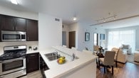 This single-floor residence offers a thoughtfully designed open-concept layout that promotes a seamless flow between its primary living spaces. The kitchen serves as a functional centerpiece, featuring dark cabinetry contrasted by a bright white countertop with a double-basin sink and a breakfast bar that accommodates casual seating. Track lighting and recessed fixtures illuminate the space efficiently, while an adjacent dining area provides a dedicated space for formal meals, complete with a glass-top table and upholstered seating. The living area extends naturally from the dining space, furnished in a soft, neutral palette that creates a calm and inviting atmosphere. Hardwood-style flooring runs throughout the main areas, adding warmth and visual continuity. The layout includes multiple bedrooms, each designed with comfort in mind, along with a well-positioned bathroom accessible from the central corridor. Throughout the residence, white walls and clean lines reinforce a modern, understated aesthetic. The overall design balances practicality with style, making efficient use of the available footprint while maintaining a sense of openness and comfort across all areas of the home.