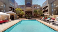 This outdoor amenity space in Dallas centers around a generously sized, sparkling swimming pool with a classic rectangular layout and clearly marked depth indicators. The pool deck is finished with neutral stone-toned tile, providing ample space for lounging and socializing under the Texas sun. Surrounding the pool, a well-appointed sundeck features rows of cushioned chaise lounges with vibrant teal and red accents, accompanied by patio umbrellas that offer shaded relief throughout the day. String lights are strung above the area, creating an inviting ambiance for evening gatherings. A white canopy cabana near the pool's edge provides a stylish focal point and additional shaded seating. The courtyard is framed by multi-story buildings with balconies, arched entryways, and warm earth-tone stucco facades, contributing to a cohesive architectural character. Mature palm trees and manicured greenery add a lush, resort-like atmosphere throughout the space. A dedicated outdoor grilling station is conveniently located nearby. Accessible, gender-specific restroom facilities are situated directly off the pool deck, ensuring convenience for all guests. The overall design of this outdoor space reflects a thoughtful blend of comfort, functionality, and upscale amenity living in the heart of Dallas.